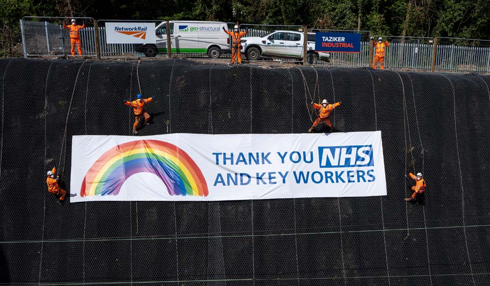 Taziker team hanging "Thank you NHS and key workers" sign over side of railway embankment.