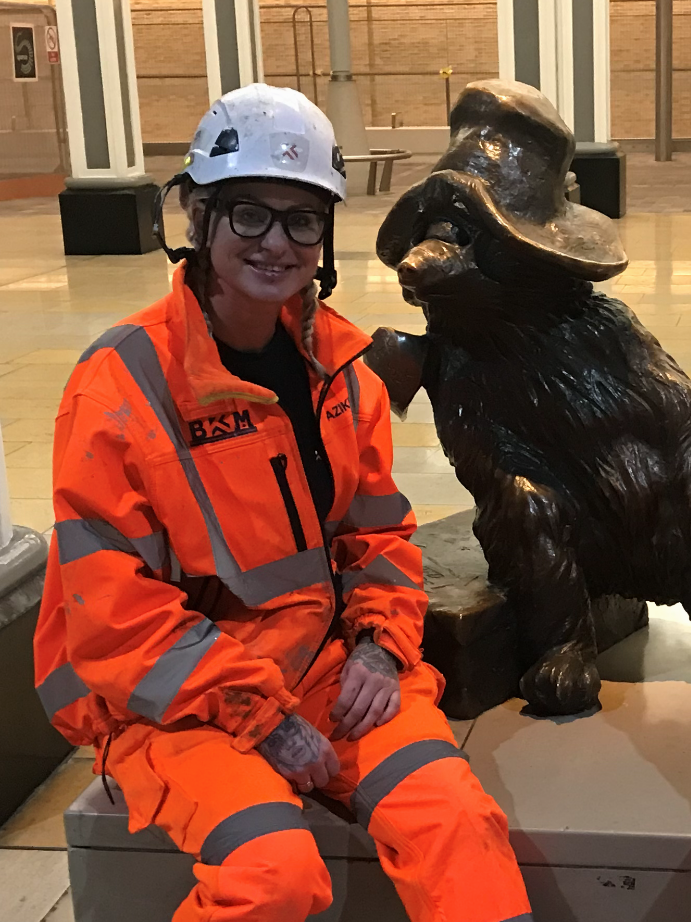 Nicola Willetts wearing PPE sat next to statue of Paddington Bear at Paddington Station.