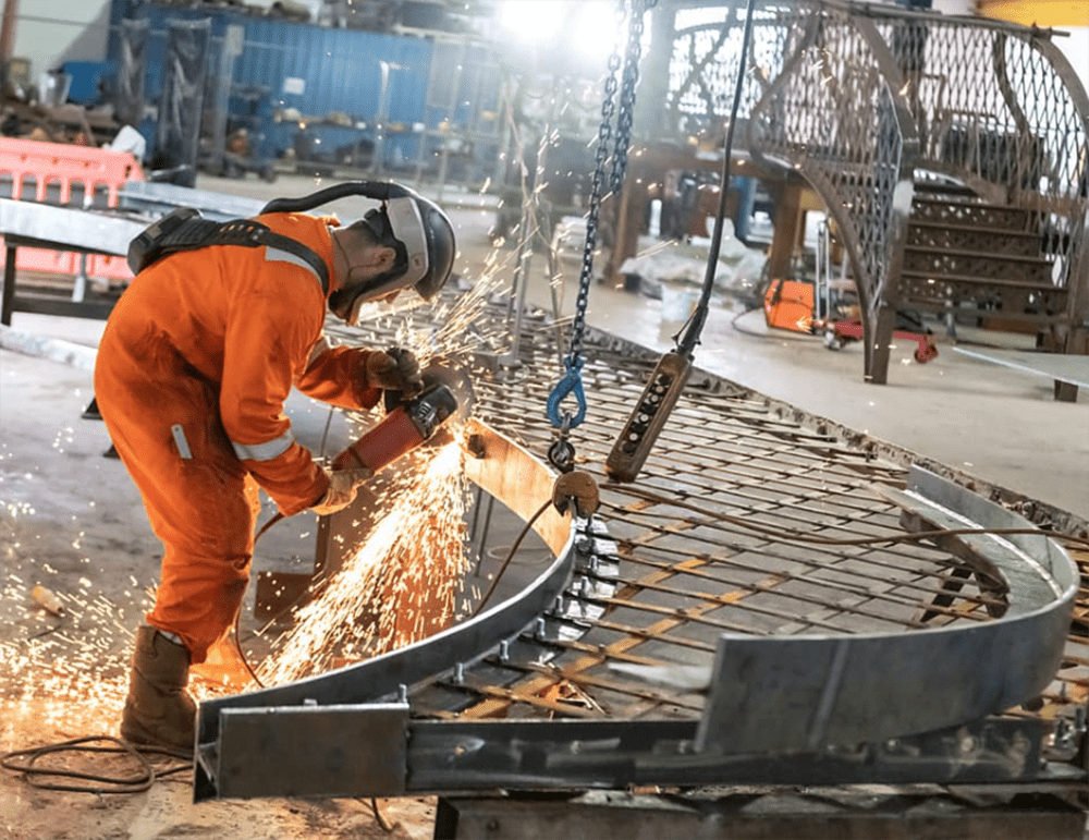 Taziker worker in orange PPE welding footbridge inside fabrication facility.